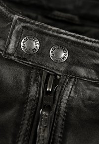 Detail einer schwarzen Lederjacke mit metallischen Druckknöpfen und "Mustang"-Branding sowie einem Reißverschluss; sichtbare Textur und Naht.