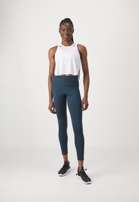 Top débardeur blanc coupé avec un col rond, associé à un legging noir de taille haute en bleu sarcelle et des chaussures de sport noires. Tissus lisses partout.