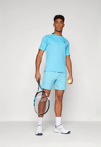 Άνδρας με ελαφριά μπλε αθλητική εμφάνιση κρατάει τένις ρακέτα στο δεξί χέρι και τένις μπάλα στο αριστερό, στέκεται μπροστά σε απλή φόντο.