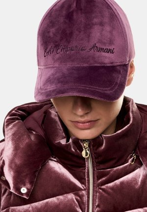 Persona che indossa un berretto di velluto viola con il logo "EA7 Emporio Armani" e una giacca imbottita abbinata, chiusa fino al collo.