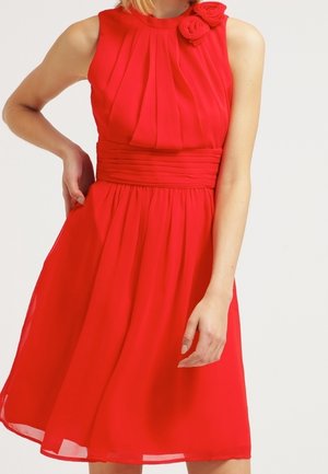 Robe rouge sans manches avec corsage plissé, large ceinture et deux décorations en tissu en forme de roses près de l'encolure.
