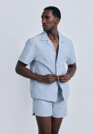 Homme portant une chemise à manches courtes rayée bleu clair et blanc avec un short assorti, boutonnant la chemise, faisant face à gauche sur un fond uni.