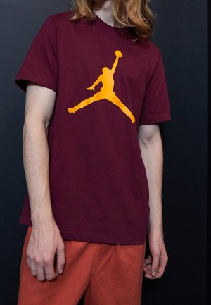 Osoba o jasnej skórze i rudych włosach nosi bordową koszulę z żółtym logo Jumpman z koszykówki oraz pomarańczowe spodnie na czarnym tle.