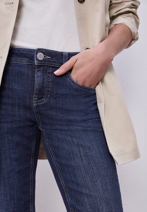 Persona che indossa jeans blu e cappotto beige, con la mano posata casualmente nella tasca anteriore dei jeans.