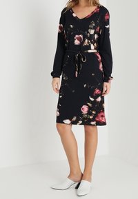 Czarna sukienka w kwiaty z długimi rękawami, dekoltem w serek, wiązana w talii, z różowymi i kremowymi kwiatami. Połączona z białymi butami na płaskiej podeszwie.
