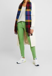 Grüne Skinny-Jeans kombiniert mit einem grauen Pullover, einem weißen langen Mantel und einem mehrfarbigen karierten Schal. Weiße Turnschuhe mit blauen und grünen Akzenten.