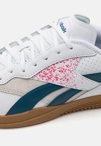 Baskets en cuir blanc avec accents turquoise, design tacheté rose, et panneau en daim gris. Elles comportent des lacets blancs et une semelle en gomme texturée.