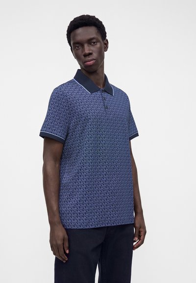 Jeune homme portant un polo bleu à motifs avec un col foncé et un pantalon foncé, debout devant un fond clair uni.