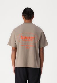 T-shirt in cotone marrone con vestibilità rilassata, caratterizzata da testo arancione sul retro: "bareen WORKSHOP LTD." e "LAVORI LIMITATI IN CORSO."