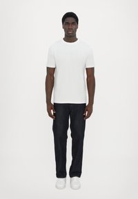 Hvit t-skjorte med korte ermer og preget logo, kombinert med mørkeblå denimjeans og hvite sneakers, stående mot en ensfarget bakgrunn.