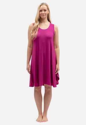 Donna in piedi scalza che indossa un abito senza maniche, ampio, color magenta con capelli biondi ondulati su una spalla.