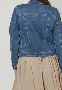 Chaqueta vaquera en azul oscuro con costuras estructuradas, corte entallado y puños abotonados, combinada con una falda de tela beige plisada.