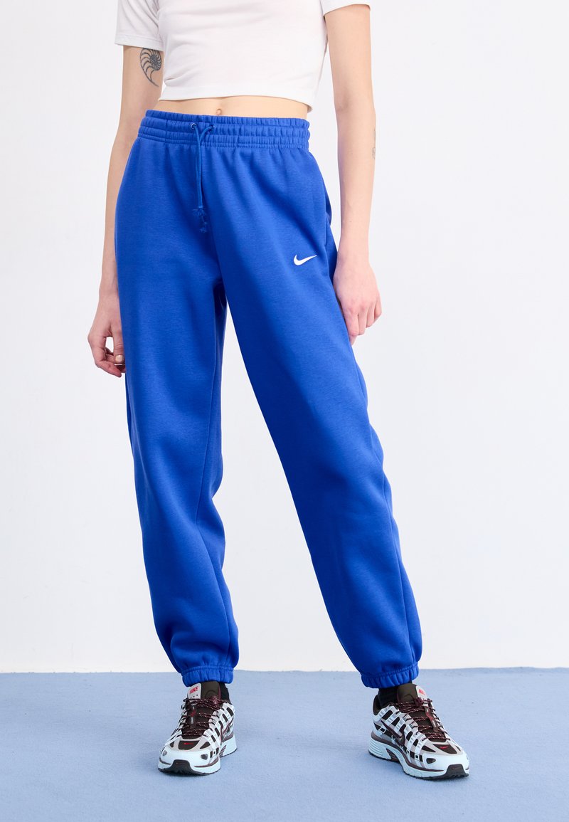 Nike blaue Sweatpants in einer entspannten Passform, mit elastischem Bund und Bündchen, versehen mit einem weißen Logo am linken Oberschenkel. Kombiniert mit gemusterten Sneakers.