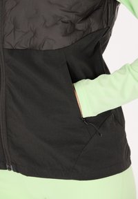 Giacca nera e verde con zip e design trapuntato, con una tasca laterale e materiale testurizzato a contrasto. Manica lunga di colore verde menta visibile.