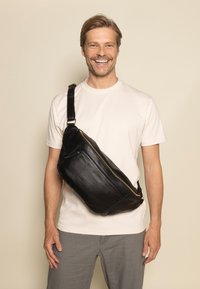 Schwarze Leder-Slingtasche mit glatter Textur, schlanker Form, goldener Reißverschluss und verstellbarem Gurt, lässig quer über einem hellen Hemd getragen.