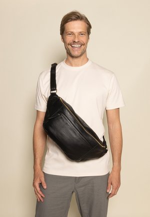 Borsa a tracolla in pelle nera con superficie liscia, forma slim, cerniera dorata e tracolla regolabile, indossata a bandoliera su una camicia chiara.