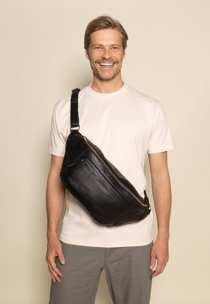 Schwarze Leder-Slingtasche mit glatter Textur, schlanker Form, goldener Reißverschluss und verstellbarem Gurt, lässig quer über einem hellen Hemd getragen.