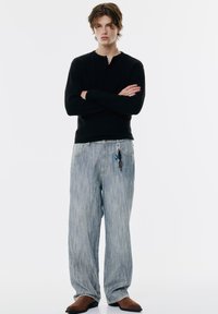 Cămașă henley cu mânecă lungă, jerseu ribbed neagră, cu o plachetă cu trei nasturi, asortată cu blugi largi, cu dungi albastre deschise și încălțăminte maro.