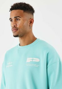 Jeune homme avec des cheveux courts et bouclés, portant des boucles d'oreilles en forme de nez et de croix, vêtu d'un sweat-shirt bleu clair avec des logos blancs imprimés sur la poitrine.