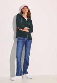 Felpa verde con zip e finitura testurizzata, abbinata a jeans a zampa d'elefante blu e sneakers bianche, completata da un cappellino viola chiaro.