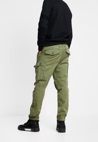 Person wearing olive green cargo pants with buttoned pockets and black sneakers, standing against a plain white background.