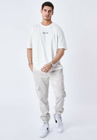 T-shirt bianca oversize con logo frontale, abbinata a pantaloni cargo grigio chiaro con tasche laterali e polsini elasticizzati, completata da sneakers bianche.