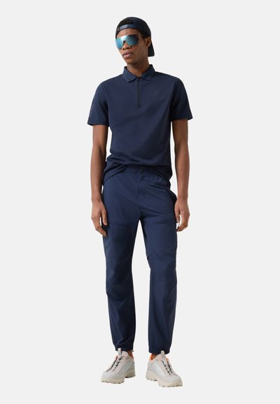 Młody mężczyzna stoi, ubrany w granatową koszulkę polo, dopasowane spodnie typu cargo, białe trampki, czapkę na odwrotnie i lustrzane okulary przeciwsłoneczne na jednolitym tle.