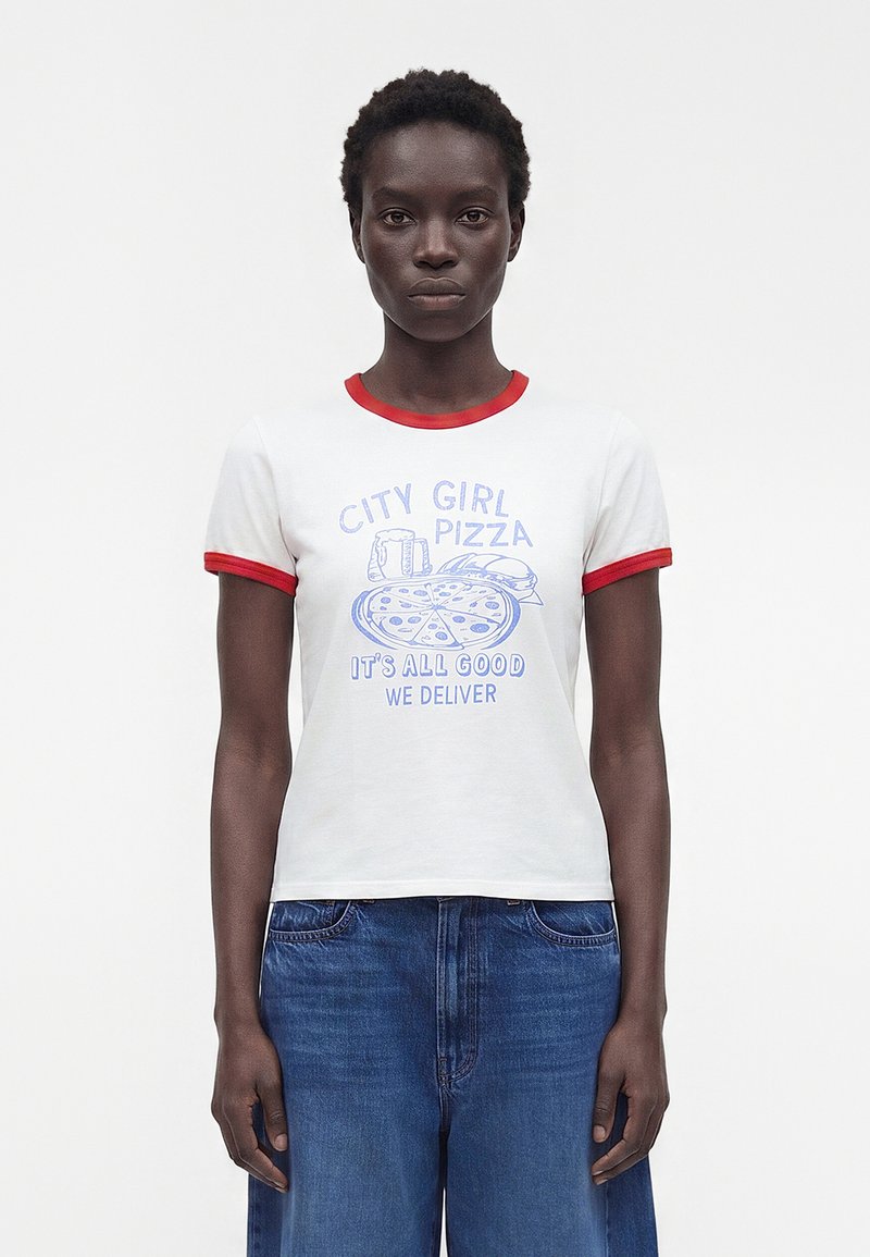 Γυναίκα που φοράει λευκό T-shirt με κόκκινη τρέσα και μπλε γραφικό κείμενο "City Girl Pizza It's All Good We Deliver" σε συνδυασμό με μπλε τζιν.