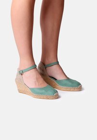 Žalios wedge espadrilės su tekstūruotu jūtos padu ir kulkšnies dirželiu. Viršus pasižymi lygaus audinio apdaila šviesiai mėtų atspalvyje.