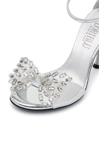 Sandal con tacco trasparente dal finish specchiato, decorata con un fiocco ornato di gemme trasparenti. Cinturino alla caviglia in argento e interno liscia.