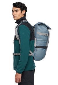 Zaino impermeabile blu con roll-top, dotato di spallacci grigi, fibbie laterali regolabili e dettagli in rete, indossato da un modello con una giacca di pile verde.