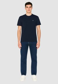 T-shirt in cotone blu navy con maniche corte e scollatura rotonda, caratterizzata da un piccolo logo sul petto, abbinata a jeans in denim scuro e sneakers bianche.