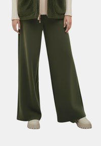 Wijd uitlopende broek in donker groene stof, gekenmerkt door een gladde textuur en een hoge taille. Gecombineerd met beige plateauzolen.