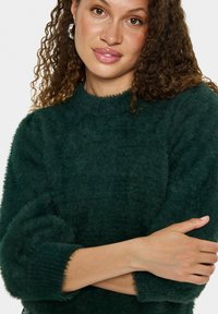 Dunkelgrüner, flauschiger Pullover mit rundem Ausschnitt und weiten Ärmeln, ausgestattet mit gerippten Bündchen. Das Material wirkt weich und strukturiert.