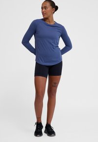 Długi rękaw, niebieska koszulka sportowa, lekki materiał, okrągły dekolt, w połączeniu z dopasowanymi czarnymi szortami i czarnymi trampkami, widoczne tatuaże na nogach.