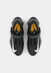 Chaussures de sport noires avec un large swoosh blanc de Nike, surface texturée, sangle ajustable et doublure intérieure orange. Convient pour l'entraînement.