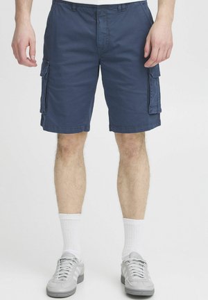 Pantaloni scurți de tip cargo navy dintr-un material ușor, cu buzunare laterale, bucle pentru curea și tiv cusut, purtați cu adidași gri și șosete albe.