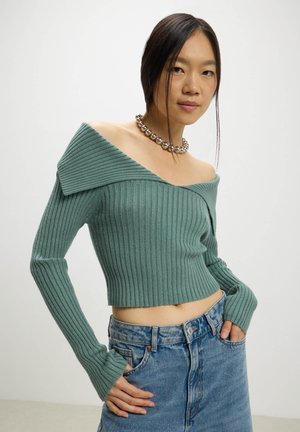 Maglione corti a coste verde con design off-shoulder, maniche lunghe e finitura testurizzata, abbinato a jeans di denim blu chiaro.