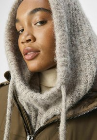 Écharpe à capuche en tricot marron clair avec une texture épaisse, superposée sur un col roulé beige, complétée par une veste vert olive.