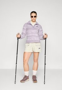 Jasnofioletowy polar z geometrycznymi wzorami, beżowe szorty cargo, szare buty trekkingowe oraz kije trekkingowe. Dodatki to okulary przeciwsłoneczne.