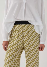 Vit skjorta med långa ärmar, kombinerad med mönstrade byxor i olivgröna vågiga ränder på en krämfärgad bakgrund, elastisk svart midja.