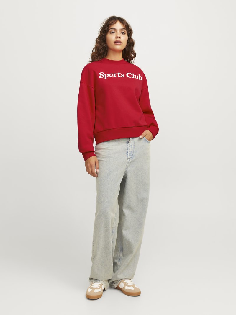 Felpa rossa con testo "Sports Club" in bianco, abbinata a jeans larghi azzurri e sneakers con dettagli beige. Design e vestibilità casual.
