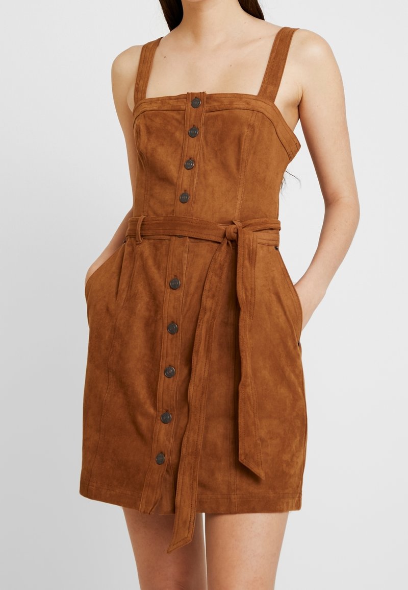 Braunes Wildlederkleid mit tailliertem Oberteil, verstellbaren Trägern, vorderen Knöpfen und einem gebundenen Taillengürtel; verfügt über Seitentaschen und eine glatte Textur.