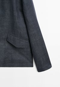 Dark gray blazer sleeve and flap pocket detail on textured fabric with a clean white background.