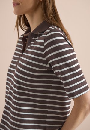 Vrouw die een bruinwit horizontaal gestreept poloshirt met korte mouwen draagt, zijaanzicht tegen een beige achtergrond.