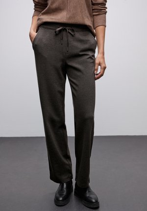 Personne portant un pull en tricot marron, un pantalon gris foncé à cordon et des bottines noires sur un sol gris et un fond gris clair.