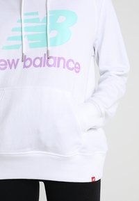 Felpa bianca New Balance con logo menta e viola, tasca laterale e cordini, indossata su pantaloni neri.
