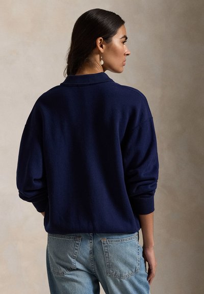 Tumepruunide juustega naine madalas kuklis, kandes mereväe sinist kampsunit ja helehalli teksapükse, küljelepoole vaadates ühevärvilise beeži tausta ees.