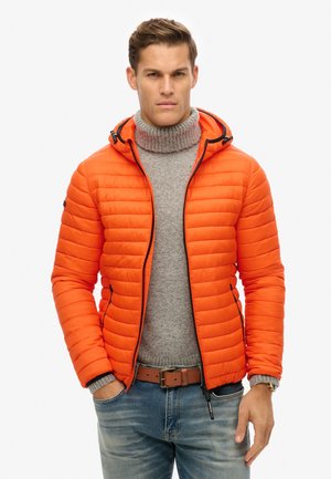 Chaqueta acolchada de color naranja brillante con acolchado horizontal, cierre de cremallera y capucha ajustable. Lleva puesta sobre un suéter de cuello alto gris claro y unos jeans azules.