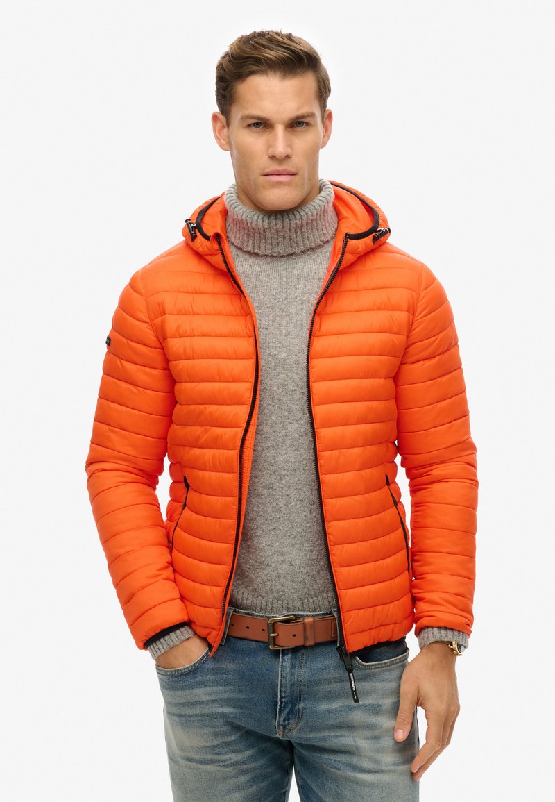 Lys orange puffjakke med vandret quilting, lynlåslukning og justerbar hætte. Båret over en lys grå højhalset trøje og blå bukser.